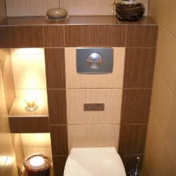 Nowoczesna łazienka z beżowo-brązowymi płytkami, podświetlanymi półkami z dekoracjami: świecą i gniazdem, oraz minimalistycznym panelem spłukującym toalety.